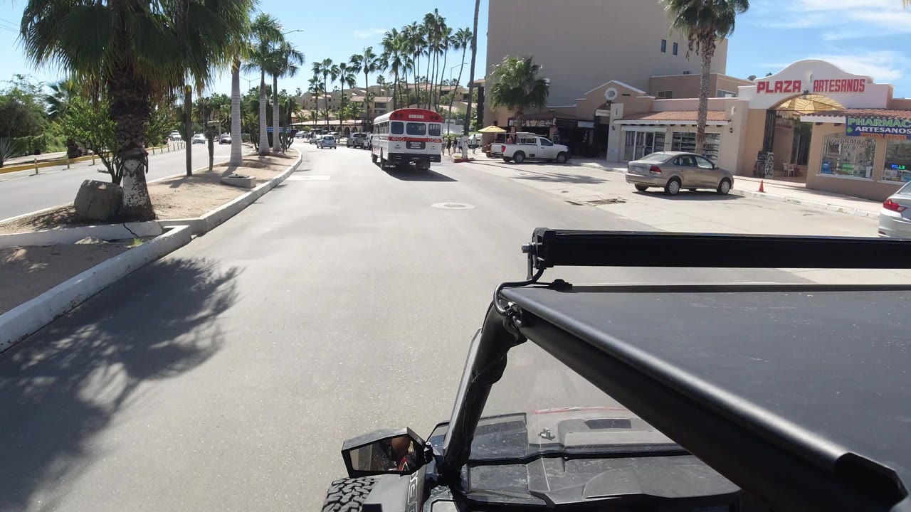 The bus remains centered ahead on the Los Cabos road while plaza storefronts, a parked silver car, palm trees, and the foreground vehicle frame fill the scene, with no clearly visible people.