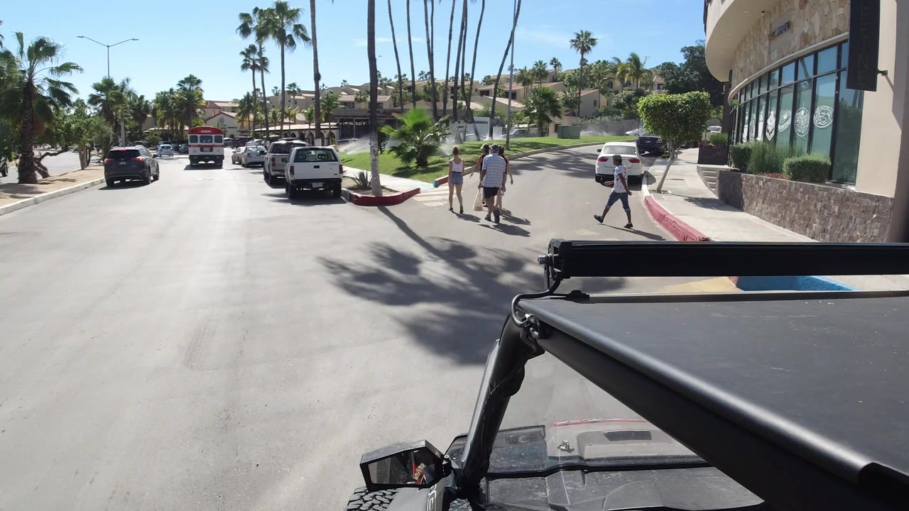 Street-level view in Los Cabos shows cars ahead, a few pedestrians near the curb, tall palms, a glass-fronted building, and the vehicle roof framing the lower edge.