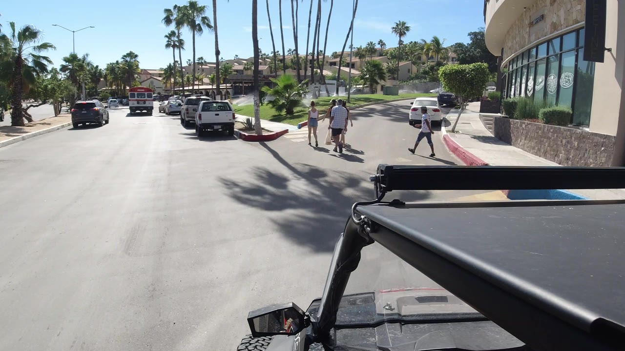 Los Cabos road scene with a white pickup and other cars ahead, pedestrians at the right curb, palm shadows on pavement, and a modern building along the sidewalk.