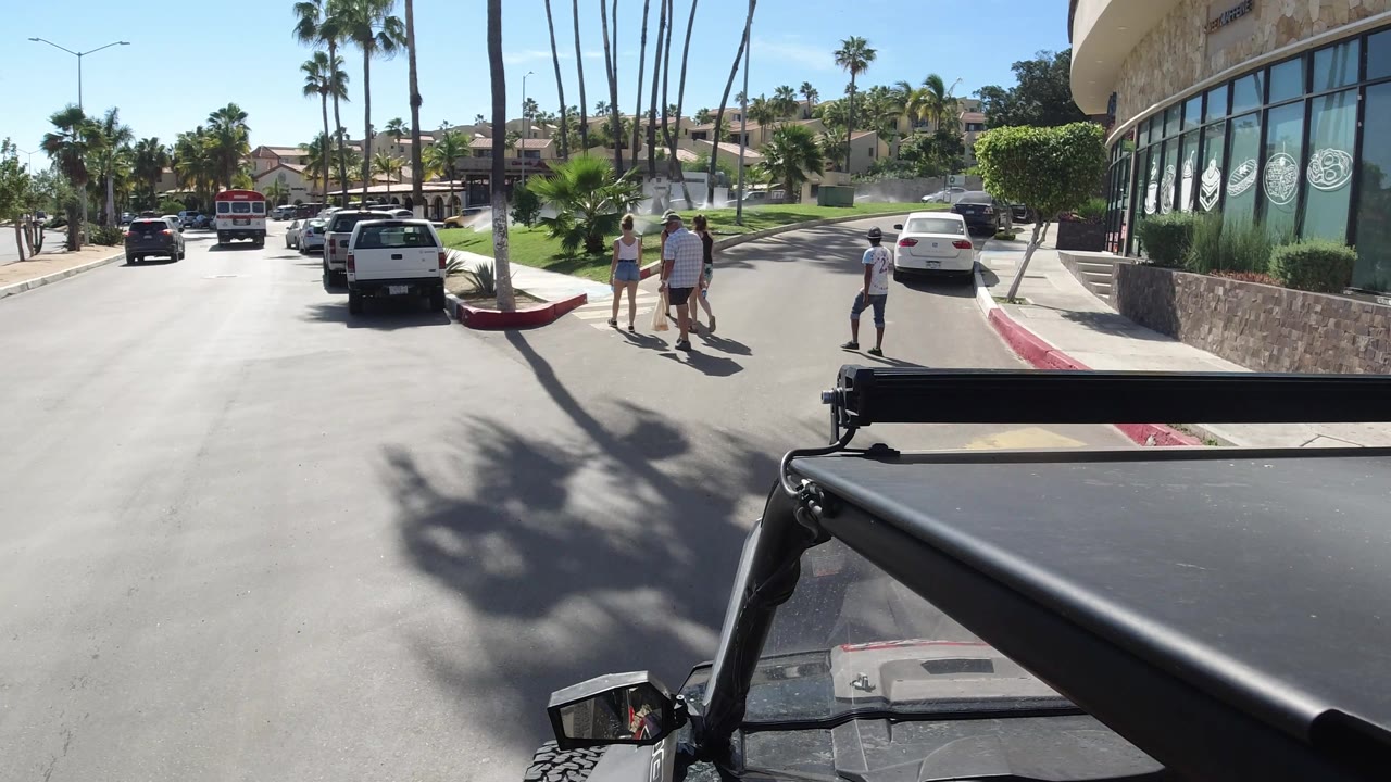 Pedestrians cross near a white car beside a curved building and palm trees.