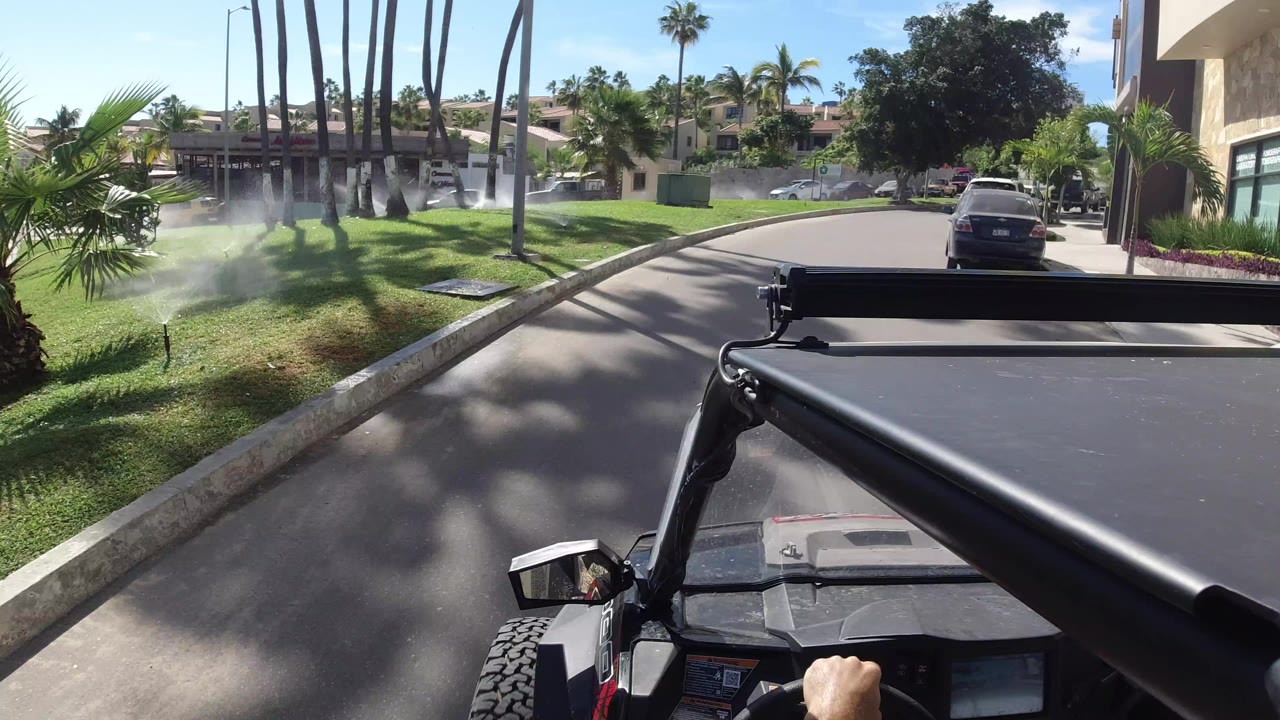 Forward-facing vehicle view in Los Cabos, Mexico, showing a sunny residential street, lawn sprinklers, tall palms, parked cars, and no visible faces.
