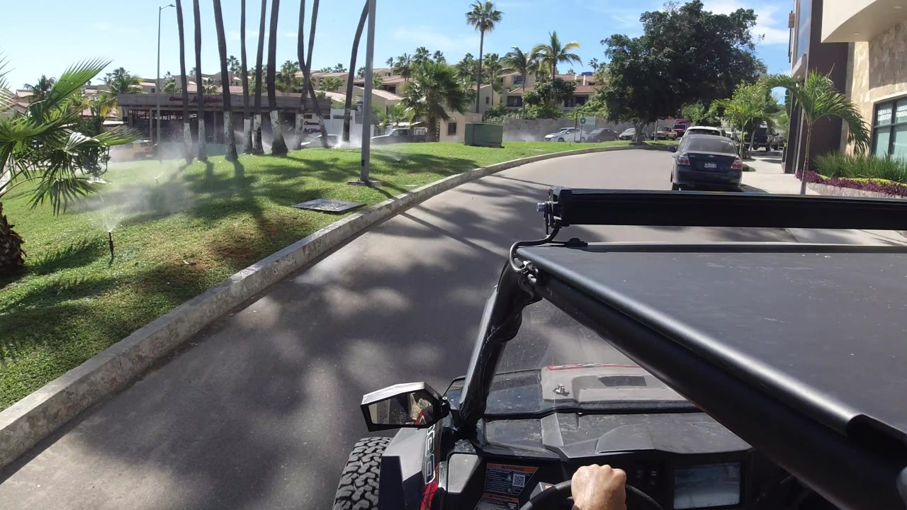 Vehicle-mounted road view in Los Cabos, Mexico, with the open vehicle foreground, palm trunks, active sprinklers, parked cars, homes, and no visible faces.