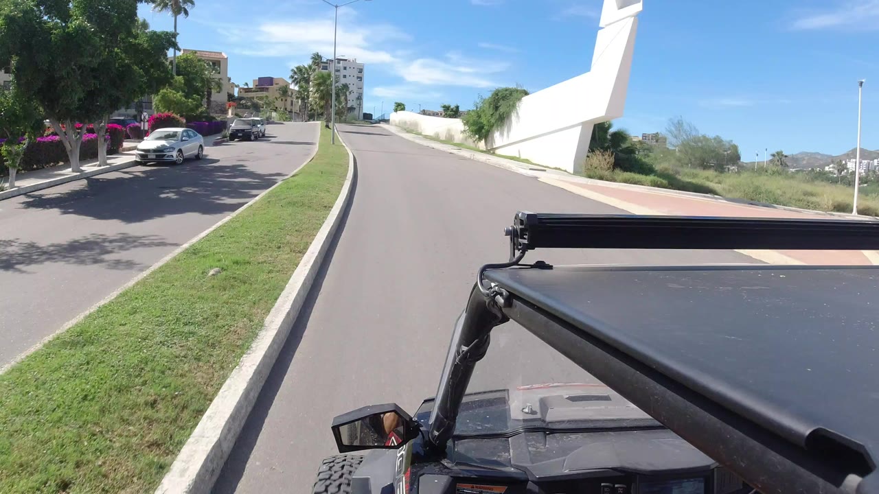 A Los Cabos street view shows a divided road, green median, parked cars, palms, and a dark vehicle roof or frame in the foreground, with no visible people.