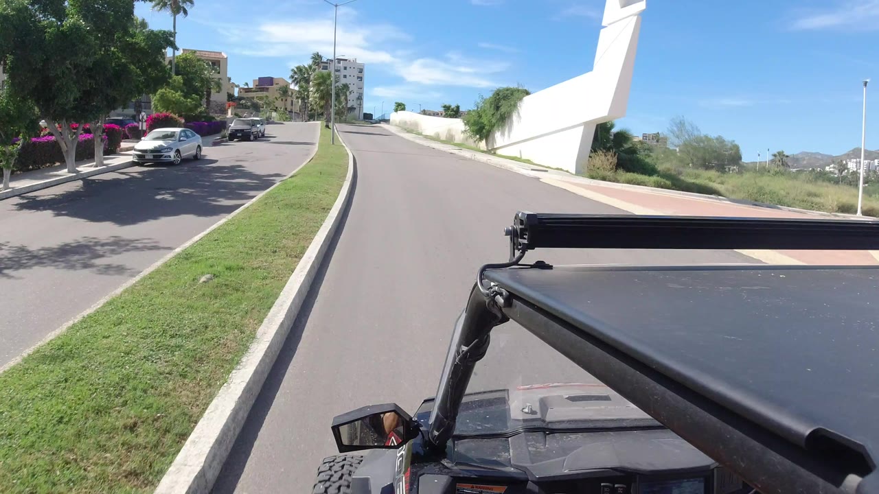 A Los Cabos roadway stretches ahead beside a white wall and grass median, with parked cars and palms to the left and no visible people.