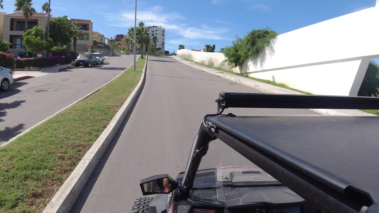 The Los Cabos street curves gently past a long white wall, green verge, palms, and low-rise buildings, with no visible people.