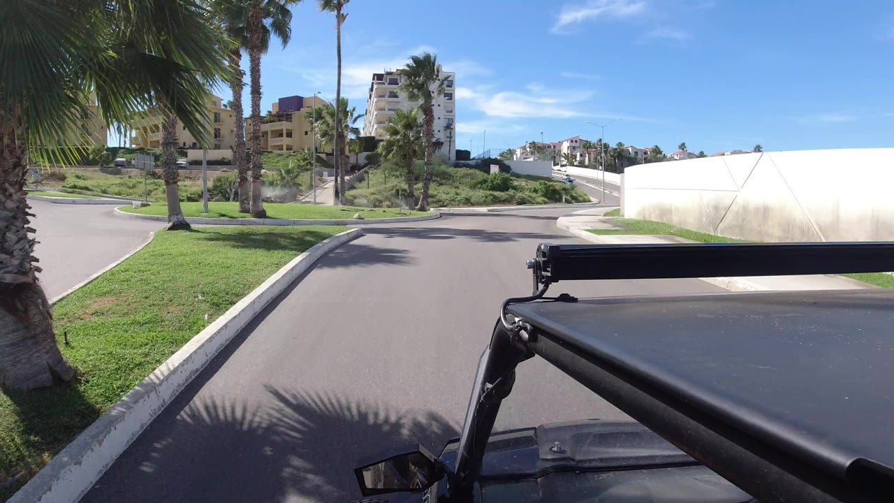 Los Cabos street view from behind a dark vehicle frame, with palms at left, a curved road ahead, white boundary walls, hillside buildings, and no visible people.