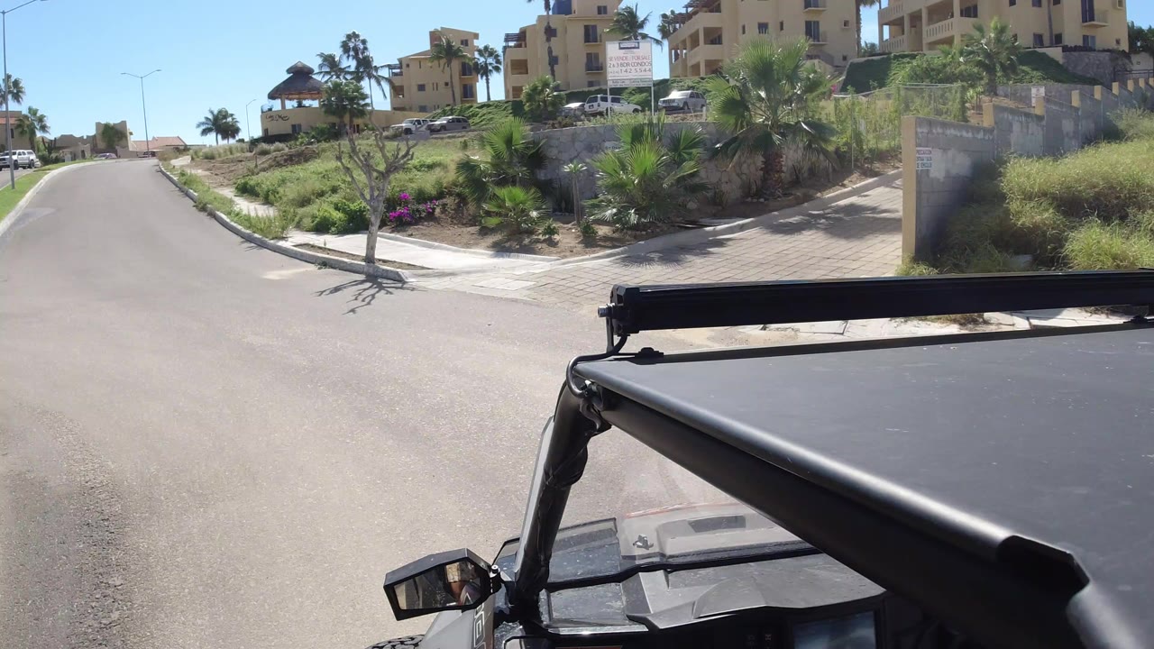 Forward view over a black Wrangler roof on a sunny Los Cabos residential street, with beige apartment buildings, palms, and no recognizable people visible.