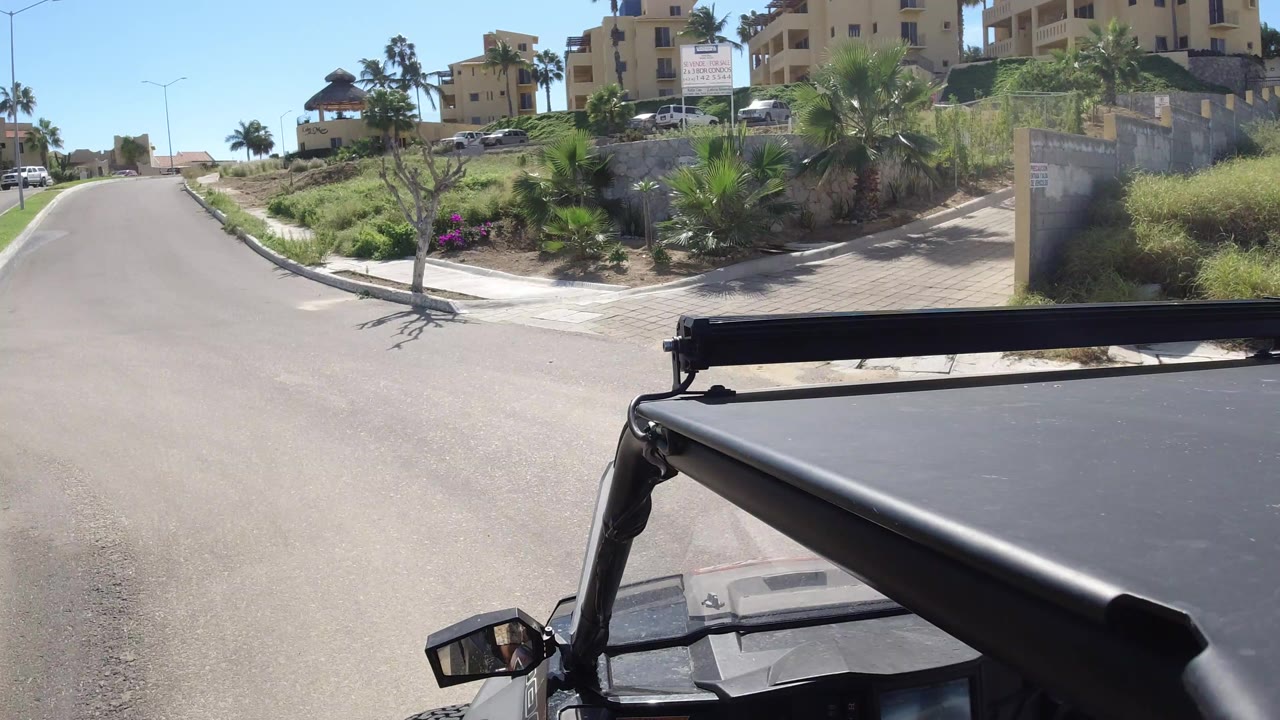 The Wrangler roof and mirror fill the lower frame as the road rises between planted medians, parked vehicles, beige buildings, and palms in Los Cabos.