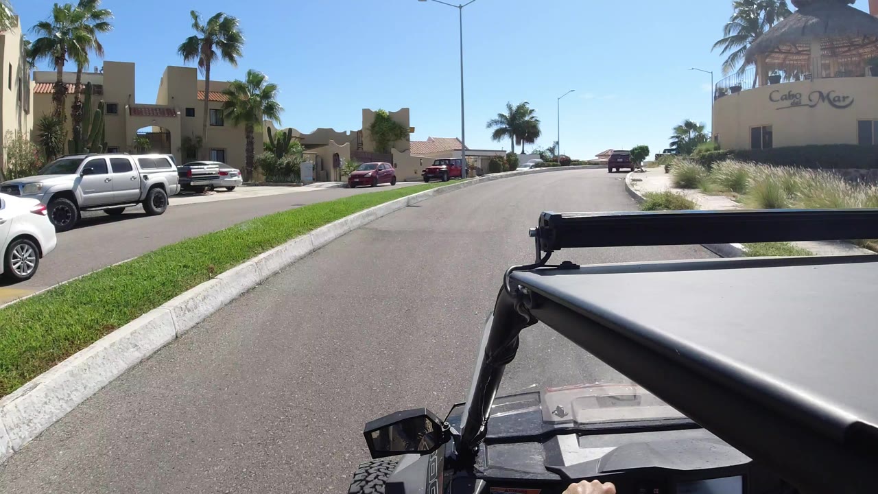 Sunny residential street with parked cars, palm trees, and open vehicle hardware in the foreground.