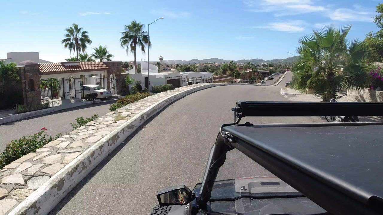 Jeep-mounted view down a sunny residential road with palms and white homes.