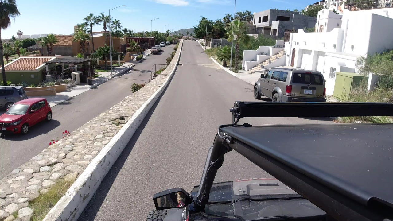 A Los Cabos residential street viewed from a moving vehicle, with a red compact car, a silver van, white homes, palms, stone curbs, and no visible people.
