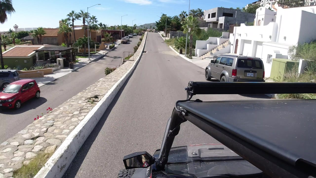 The vehicle advances along a quiet Los Cabos street, with a red car near the curb, a silver van by a white home, palms, low walls, and no visible people.