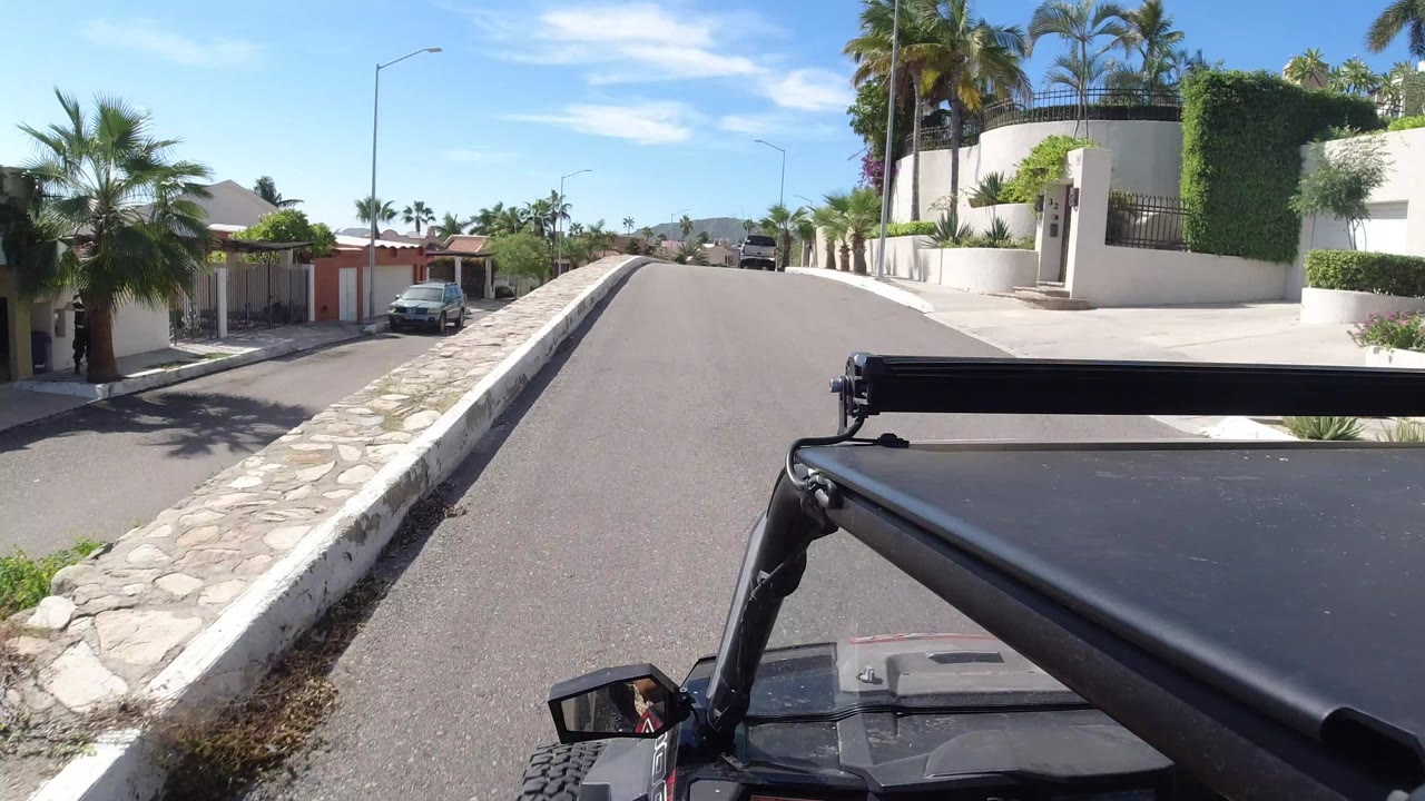 High vehicle-mounted view in Los Cabos looking up a quiet paved residential street with palms, white walls, a parked car, distant hills, and no visible people.