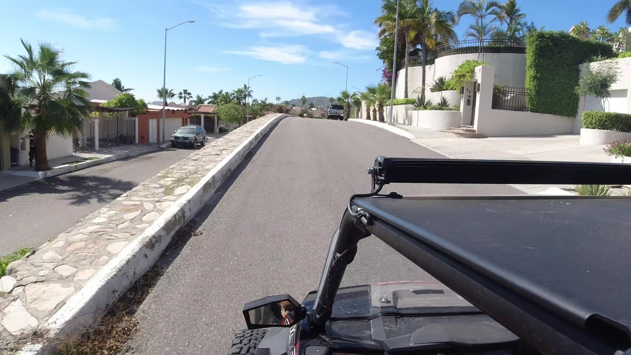 Los Cabos street view from a high mounted camera, showing the black vehicle hardware in front, white boundary walls, palm landscaping, tiled roofs, and no visible people.