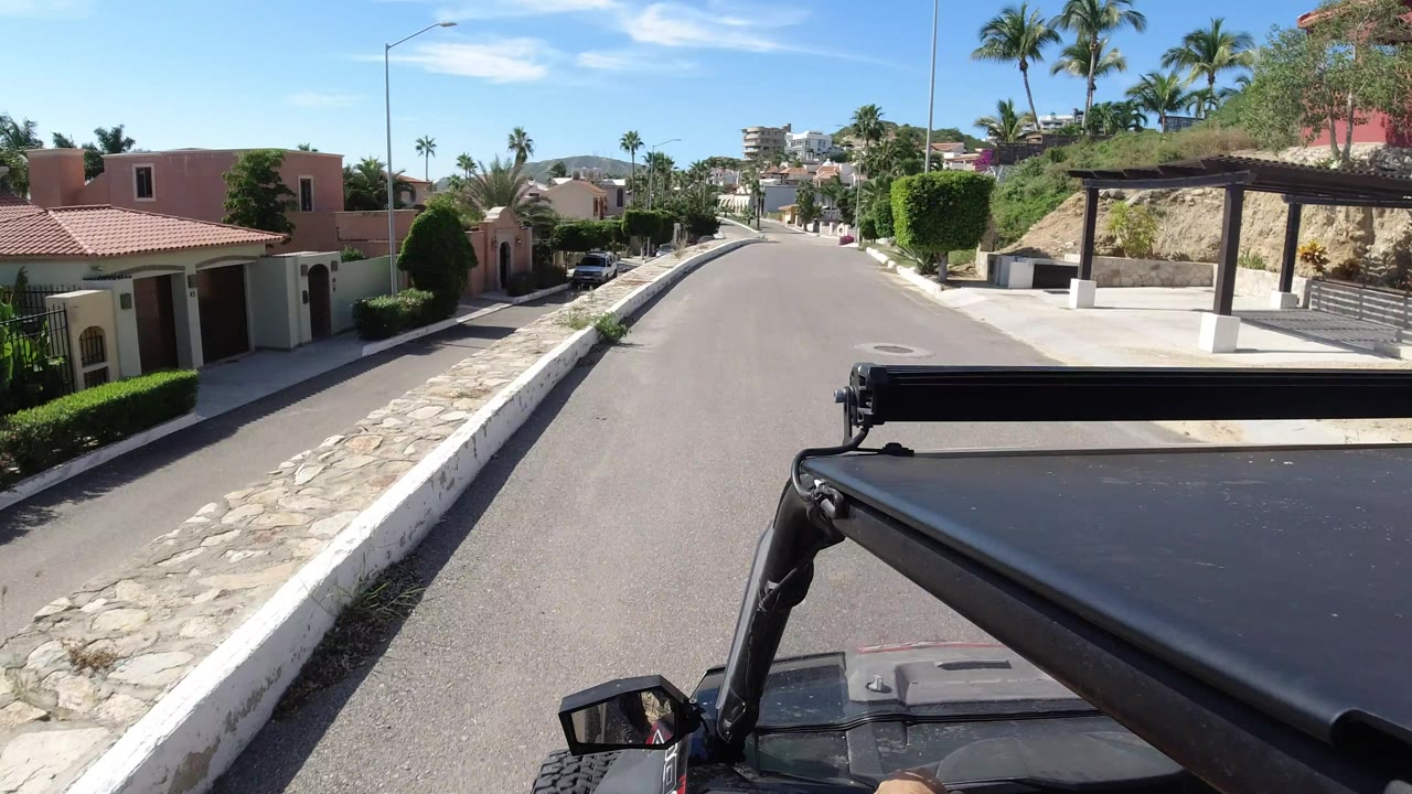 The Los Cabos road continues ahead from above the black Wrangler roof, with pink stucco walls, trimmed greenery, palms, and no visible people.