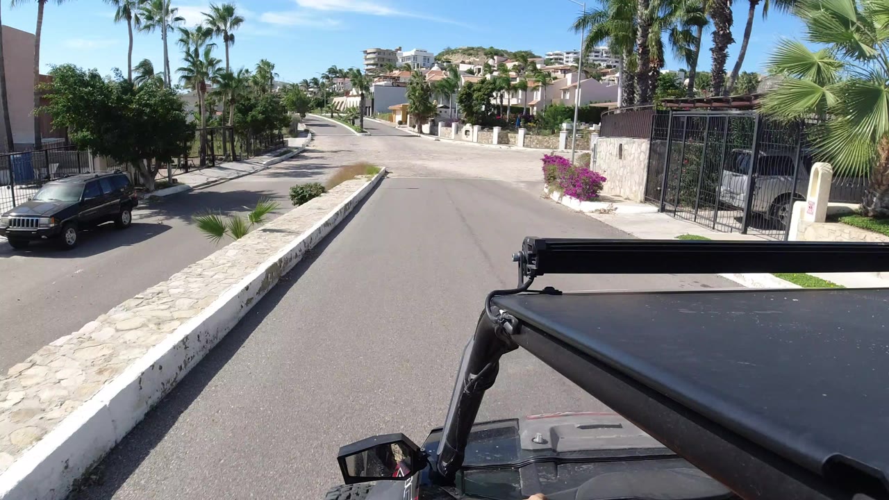 Forward view from an open Jeep in Los Cabos, Mexico, looking down a sunny paved residential street with palm trees, walled homes, a parked dark SUV, and no visible faces.