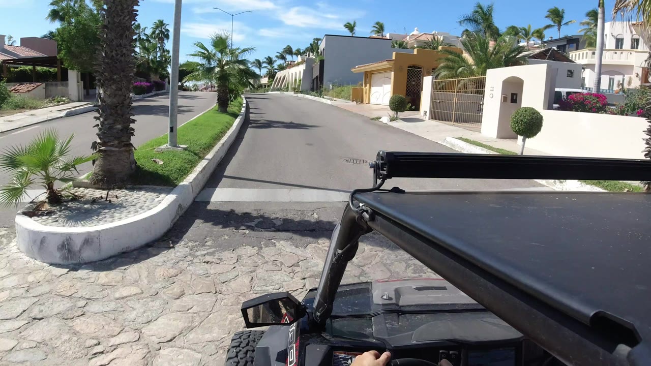The vehicle viewpoint continues along a Los Cabos residential street with a palm planter on the left, walled houses on both sides, a streetlight, and no visible people.