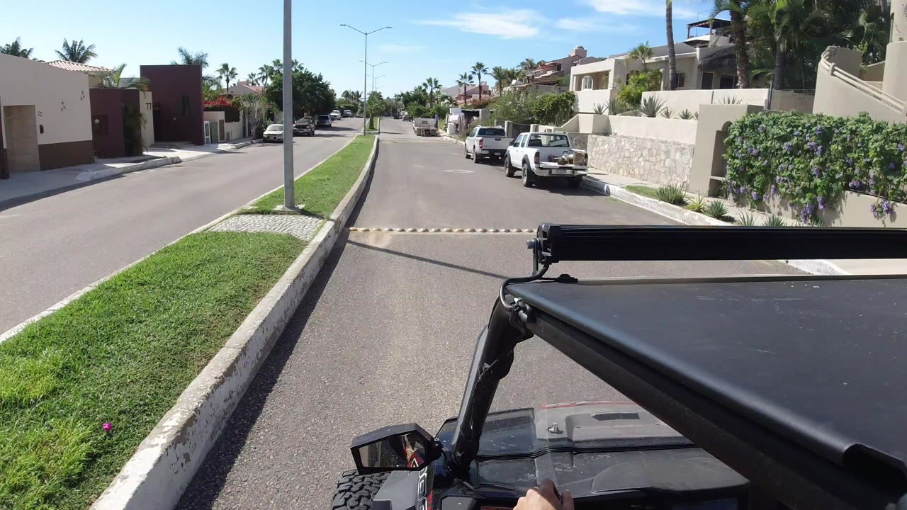 Opening Los Cabos road view from an open vehicle, with a grassy median, cream walls, parked white vehicles, palm trees, and no visible faces.