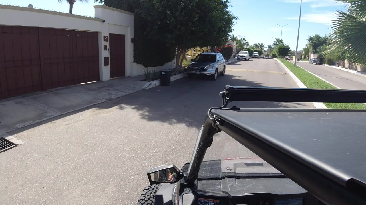 At 0:00 in Los Cabos, Mexico, the vehicle foreground faces a red driveway gate, white wall, parked cars, trees, and a sunny residential street with no visible people.