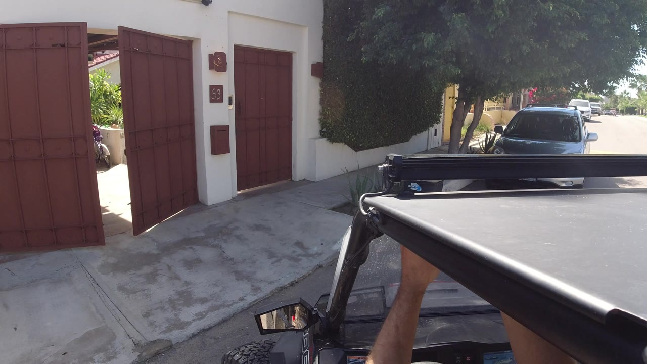Mounted Jeep view in Los Cabos with the red gate open wider, a dark car ahead under trees, cream residential walls, and a forearm beside the vehicle frame.
