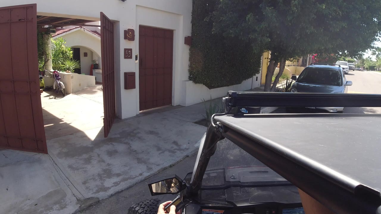 At 0:00 in Los Cabos, Mexico, a red-brown gate stands open beside a white stucco home entrance, with a dark parked vehicle and shaded trees visible beyond.