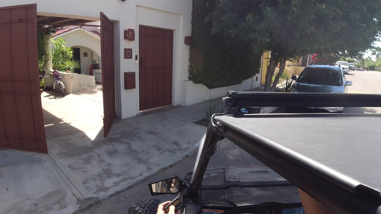 At 0:01 in Los Cabos, Mexico, the open gate frames a driveway and residential street, with a dark car parked under trees and strong sunlight hitting the pavement.