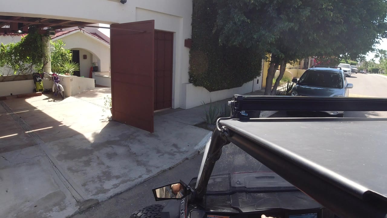 At the start of the Los Cabos clip, an open red gate frames a stucco residential entry while the vehicle roof and roll bar fill the foreground, with no visible people.
