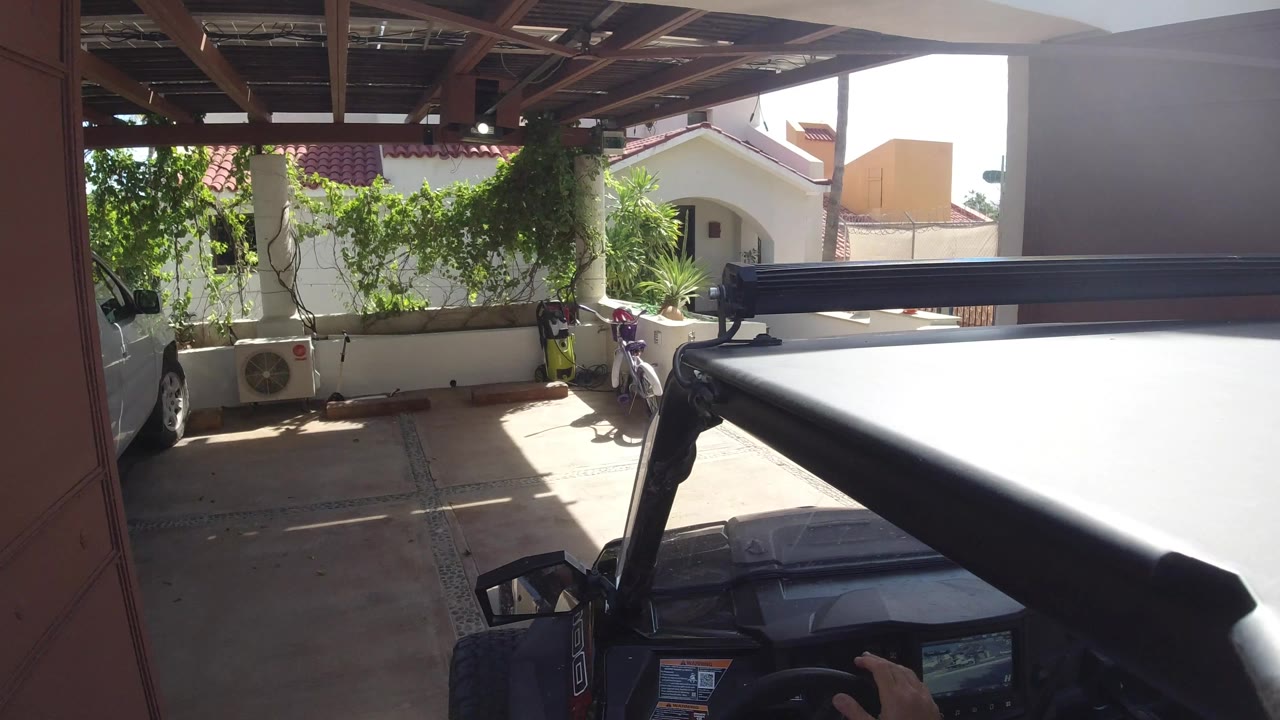 White vehicle positioned along a Los Cabos carport, with a dark foreground roofline, leafy vines, air-conditioning units, and a hand visible at the bottom of the frame.