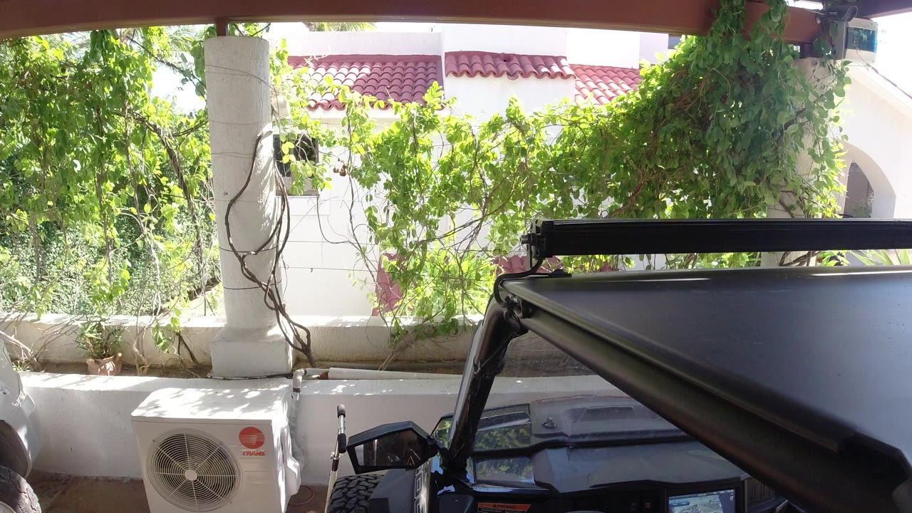 At 0:00, a black Wrangler sits partly in frame inside a shaded garage in Los Cabos, Mexico, facing a white building with terracotta roof tiles, green vines, and no visible people.