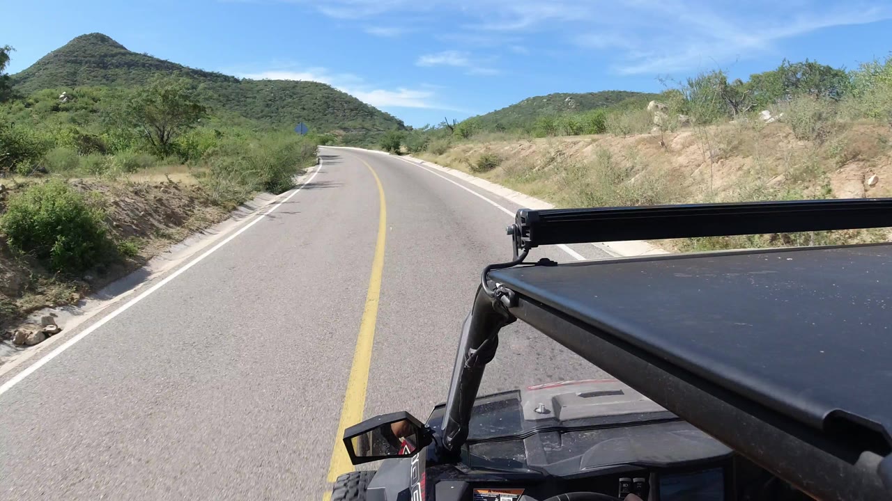 A left-window Wrangler view follows an empty paved road toward rounded Los Cabos hills, with dry shoulder cuts, roadside shrubs, and no visible people.