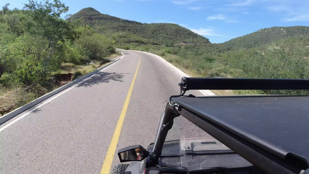 Forward-facing road POV in Los Cabos, Mexico, with a yellow centerline, green roadside brush, low hills ahead, a blue sky, and no visible people.