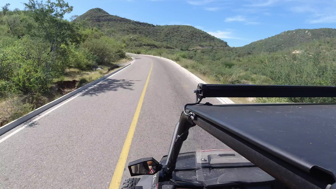 The vehicle POV continues along a sunlit Los Cabos road, with the lane bending gently ahead, dense brush on both sides, hills in the distance, and no visible people.