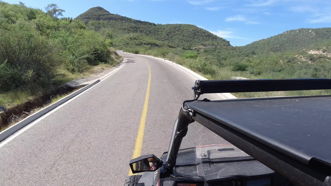 An empty two-lane road in Los Cabos runs past roadside vegetation, with a darker hill rising ahead, the vehicle frame at lower right, and no visible people.