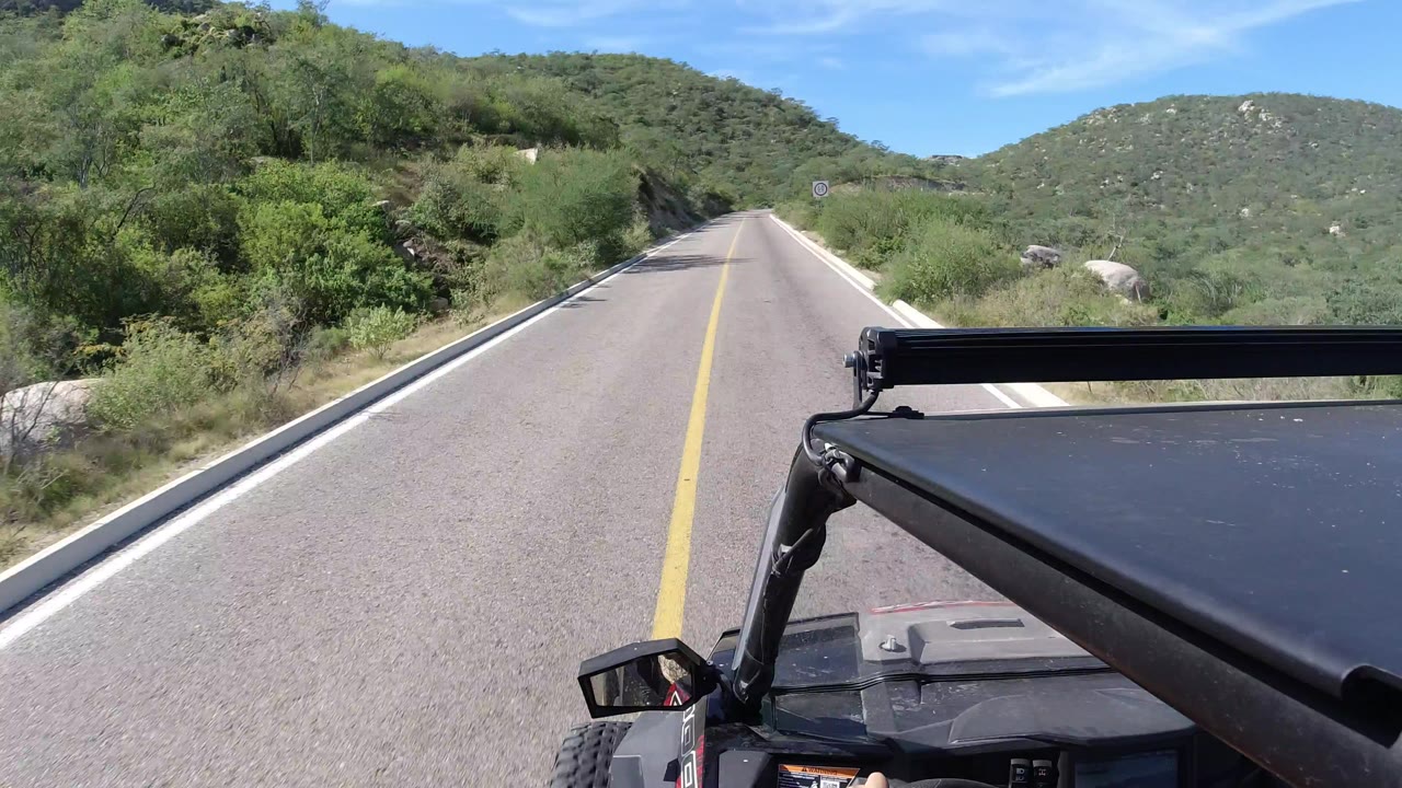 Forward view over a black vehicle roof or roll bar on a paved road through green hills in Los Cabos, Mexico, with no visible people.