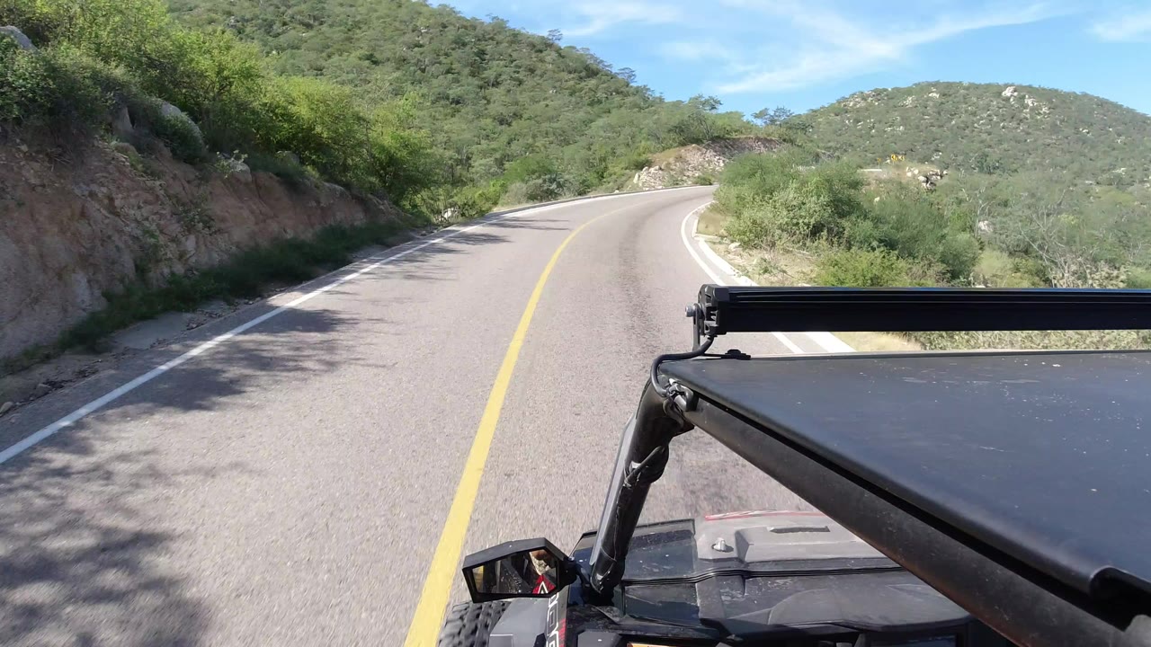 Forward view over a Wrangler hood on a curving Los Cabos road, with scrubby green hills, blue sky, and no visible people.