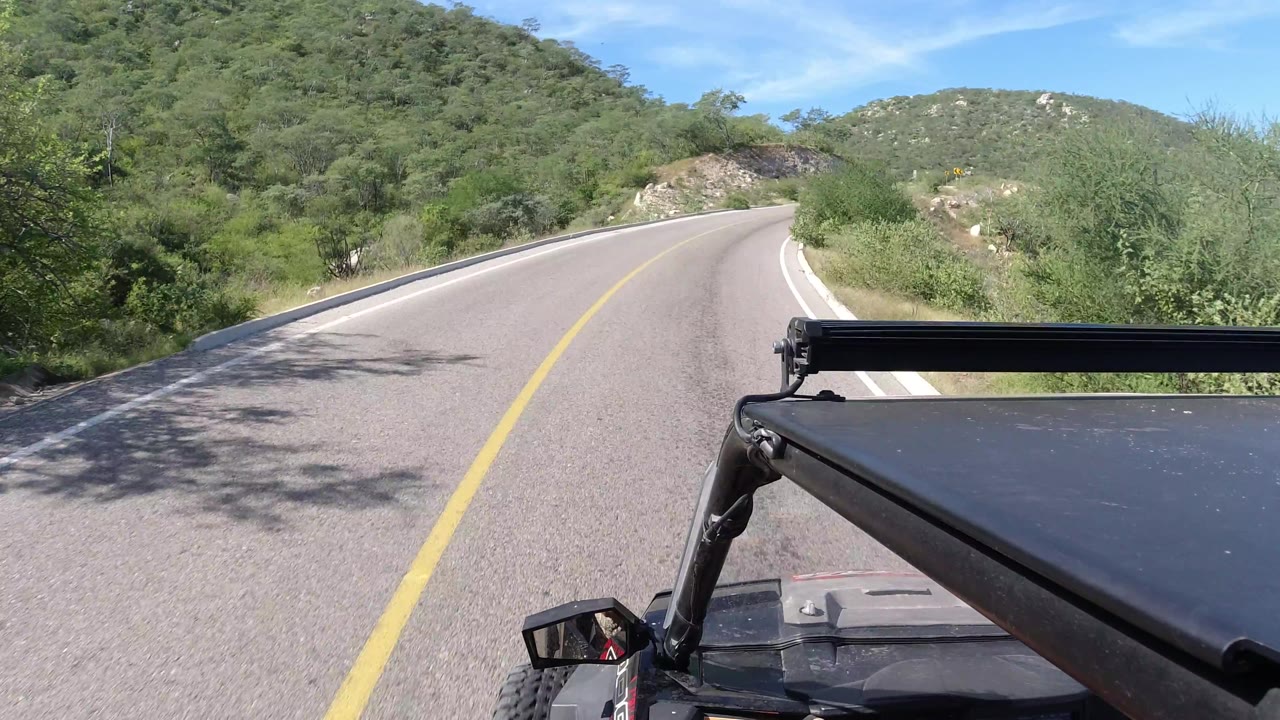 Road curve with hillside vegetation and vehicle hood at right.