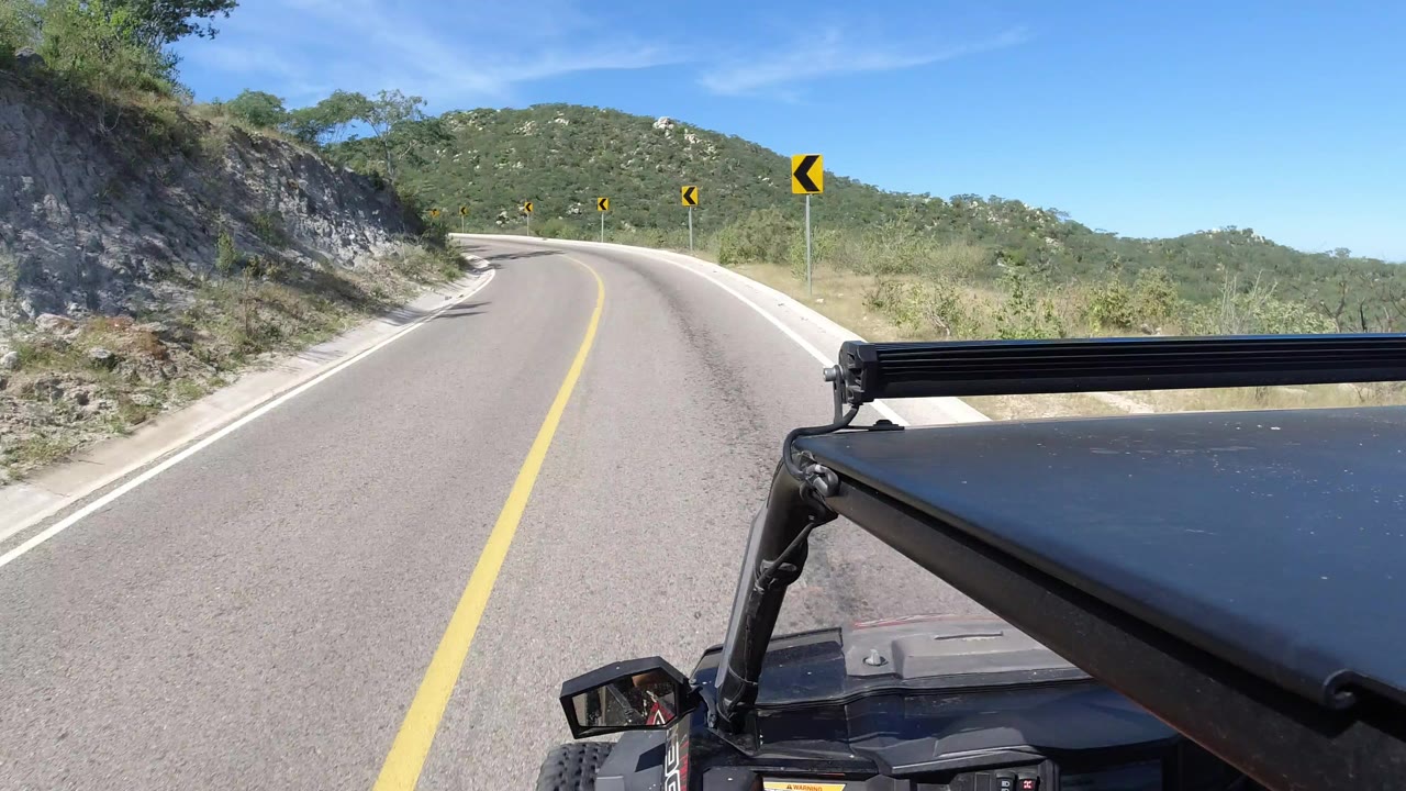 Start of the Los Cabos, Mexico clip showing a paved road bending left through green hills, with yellow curve signs ahead and no visible faces.