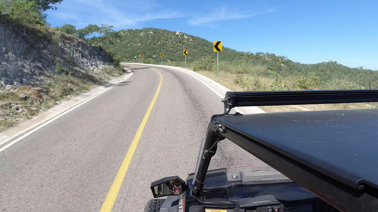 A second Los Cabos road POV frame shows the vehicle closer to the left bend, with roadside rocks, scrub plants, arrow signs, and no visible faces.