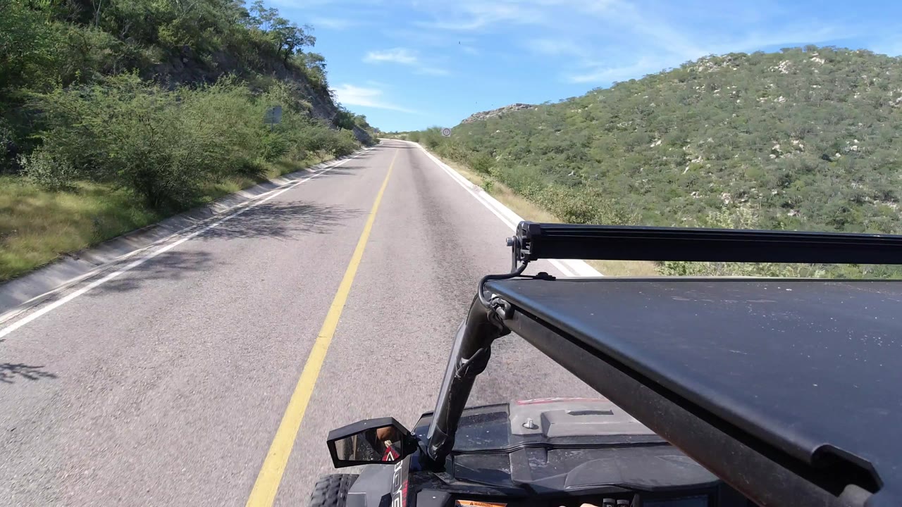 A Los Cabos road continues ahead between rocky green slopes, with the vehicle roof and black rack filling the lower right foreground and no visible people.