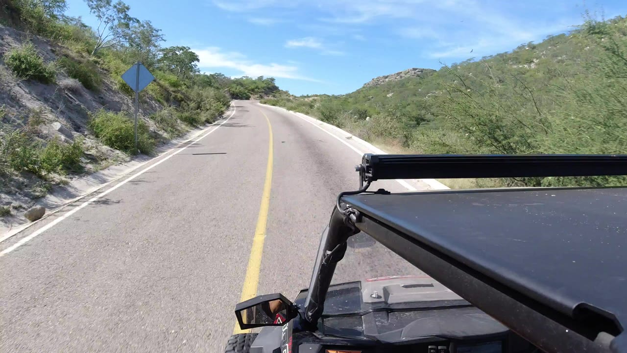 Start of the Los Cabos, Mexico clip showing an empty paved road ahead, a yellow centerline, roadside brush, a sign shape at left, black Wrangler hardware in the foreground, and no visible people.