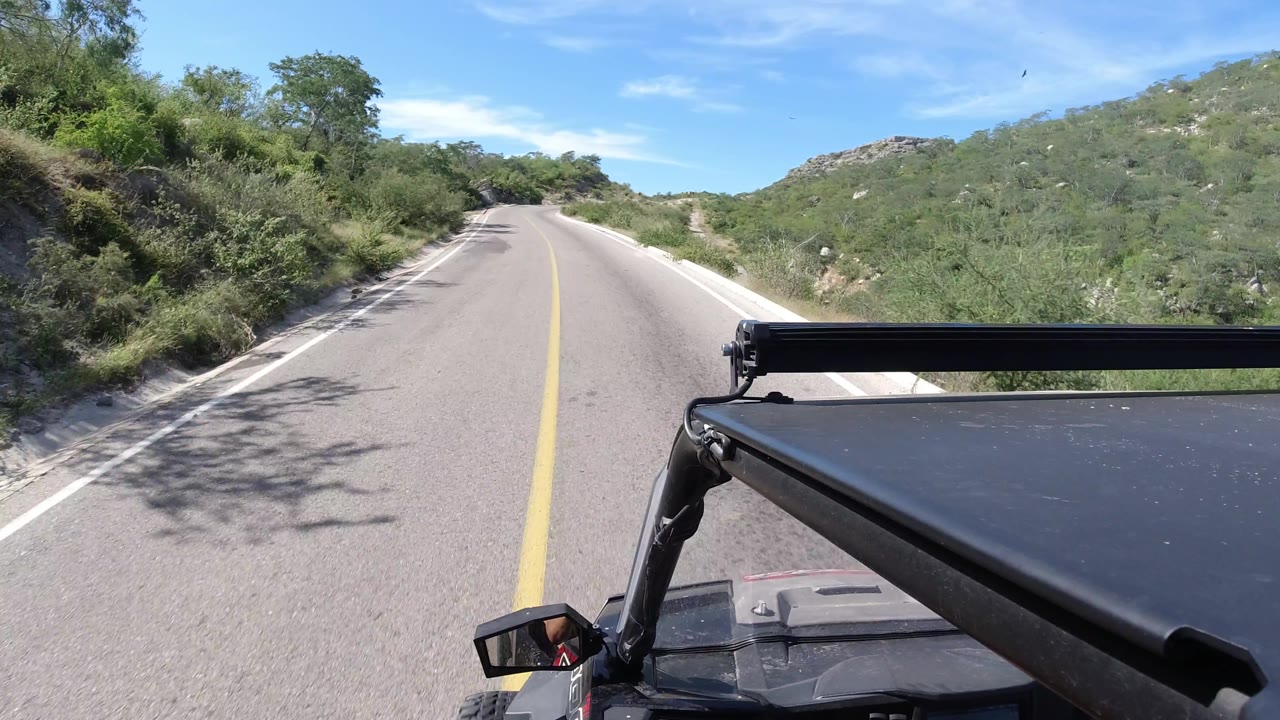 At 0:02, the Los Cabos road begins a clearer right bend, with tree shadows crossing the pavement, black Wrangler hardware at lower right, rocky hills beyond the brush, and no visible people.