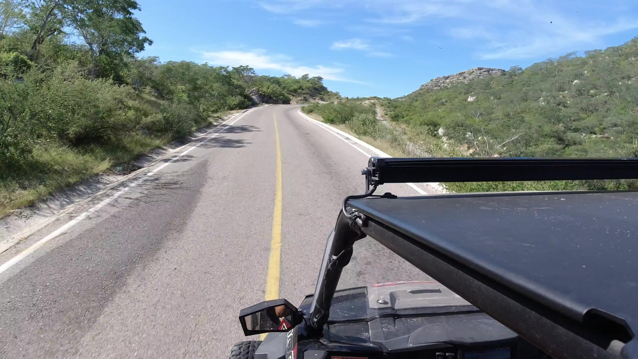 At 0:03, the empty Los Cabos highway runs ahead between dense roadside trees and low rocky hills, with the black Wrangler hood and roll bar still visible in the foreground and no visible people.