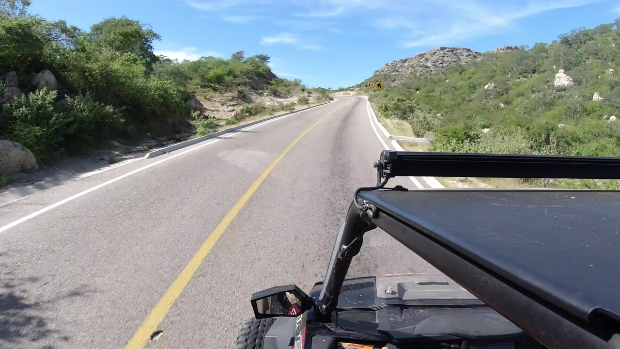 Start of a Los Cabos, Mexico road view showing an empty paved lane, yellow center line, shrubs on both sides, rocky hills ahead, blue sky, and no visible people.