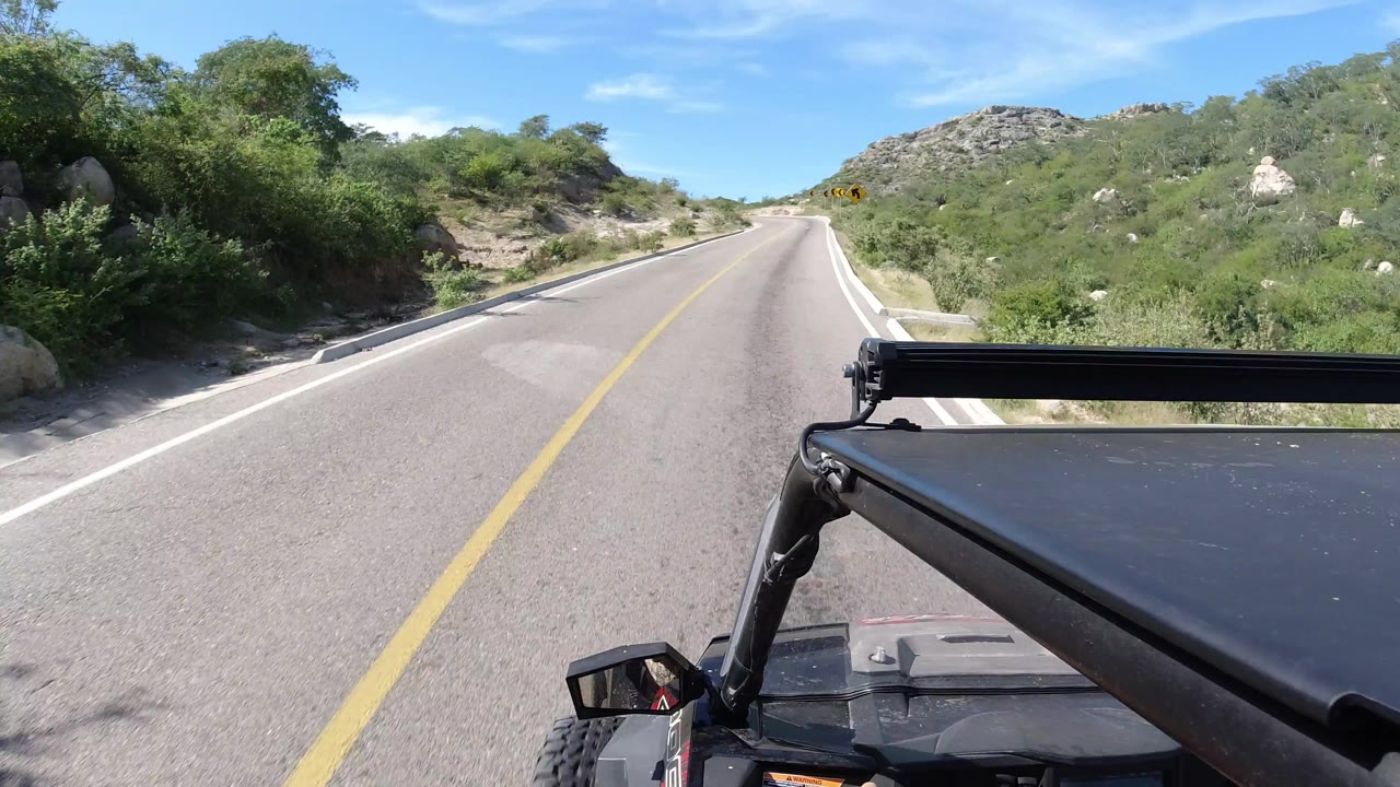 Los Cabos, Mexico road frame with the yellow line leading toward low rocky hills, dense green roadside brush, black vehicle hardware at lower right, and no visible people.