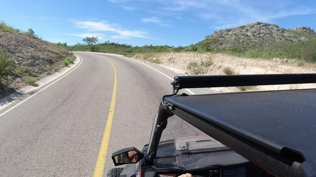 One second in, the Los Cabos road curve remains ahead with the black vehicle roof and support bar covering the lower right foreground, dry brush on both shoulders, and no visible faces.