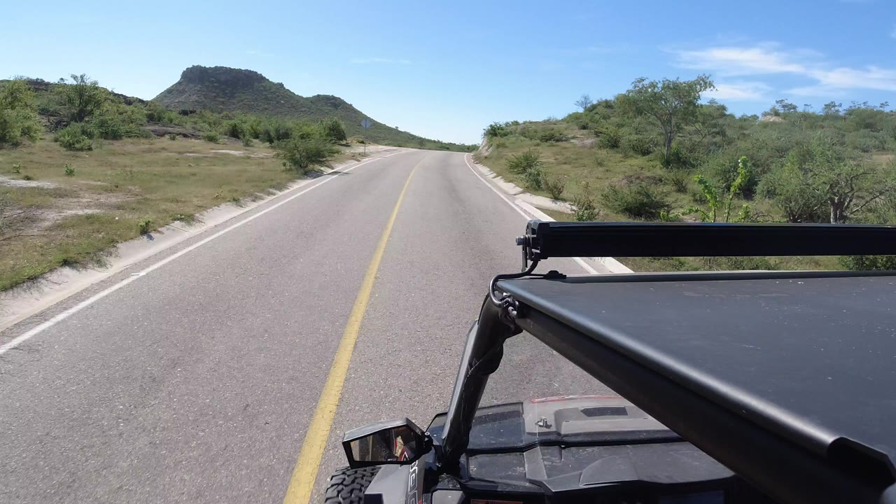 Forward view over a black Jeep roof on a paved road toward low desert hills.