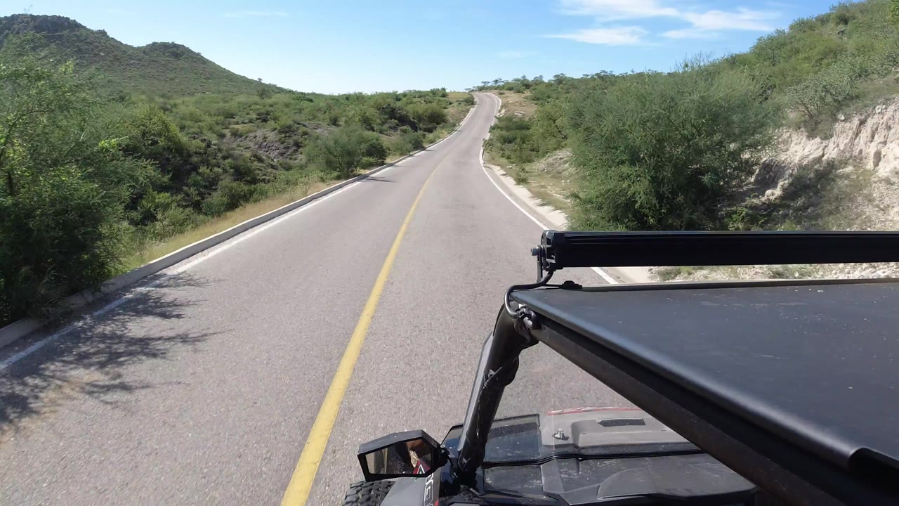The Los Cabos road continues ahead under bright sun, with the vehicle roof frame at lower right and dense green shrubs along both shoulders.