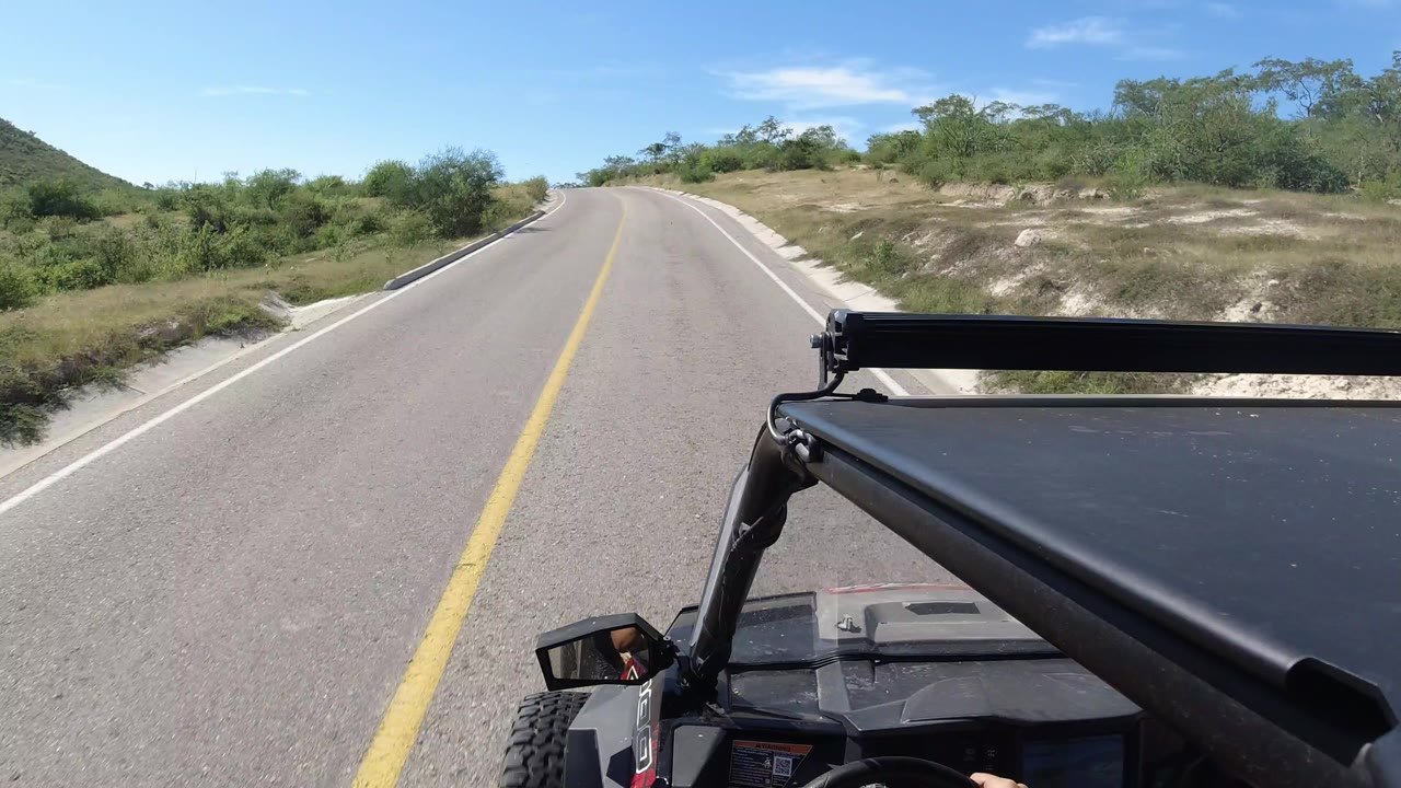 Forward view from a black open vehicle on a paved two-way highway in Los Cabos, Mexico, with dry roadside brush, low hills, bright blue sky, and no visible people.