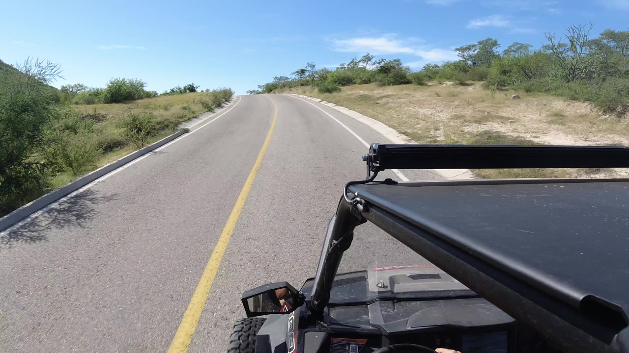 A curving paved highway in Los Cabos cuts through dry grass, shrubs, and low brush, with a black open vehicle in the foreground and no visible people.
