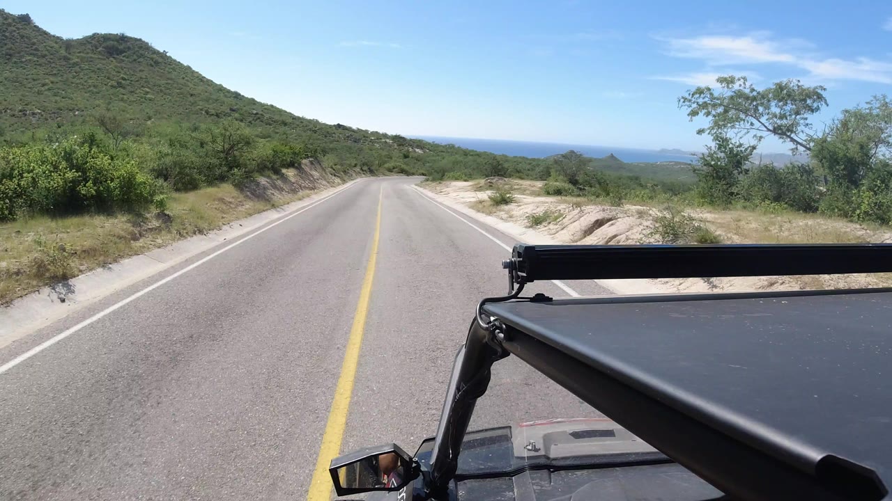 Vehicle-mounted view down a Los Cabos desert road, showing the light bar in the foreground, hillside brush on the left, distant horizon ahead, and no visible people.